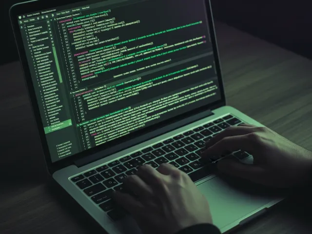 Duas mãos digitando em um teclado de laptop, com código-fonte visível na tela em um ambiente de desenvolvimento, ilustrando a rotina de um desenvolvedor de aplicativo.