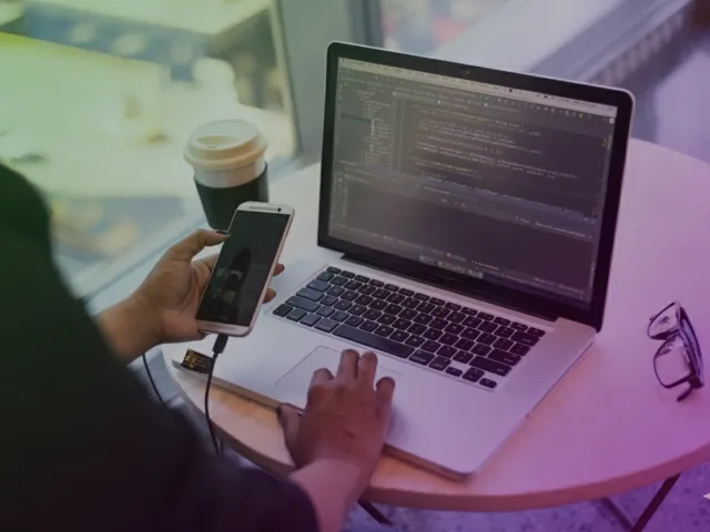 Profissional concentrado em mesa redonda, usando notebook com código e smartphone conectado por cabo, com copo de café e óculos sobre a mesa, em ambiente de trabalho com filtro em tons de verde e roxo escuro.